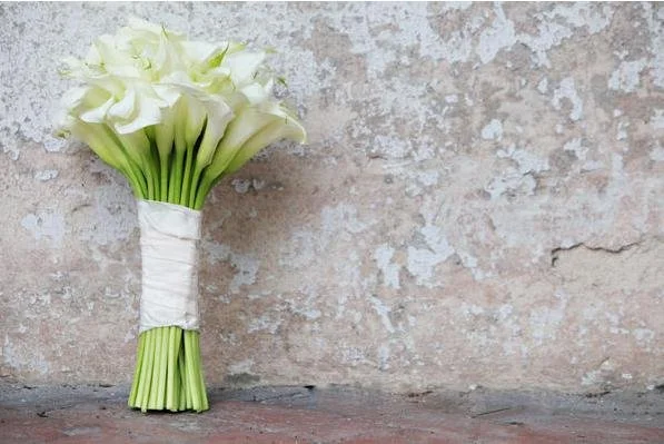 A bouquet of white calla lilies wrapped with white paper and placed on a concrete surface against a textured wall.