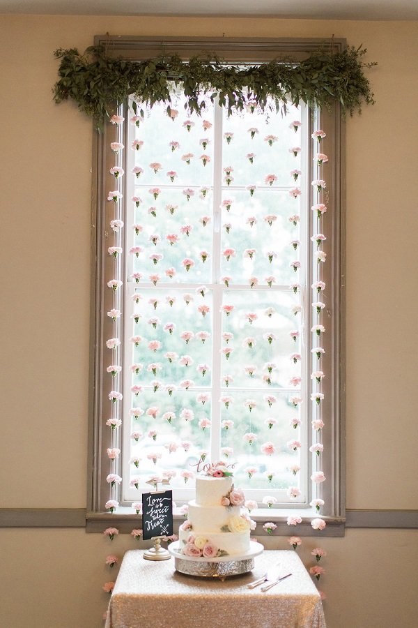 A window decorated with hanging pink and white flowers and green foliage at the top. A small table with a white wedding cake adorned with pink roses is positioned in front of the window. The cake has a chalkboard sign next to it and a 'Love' topper.