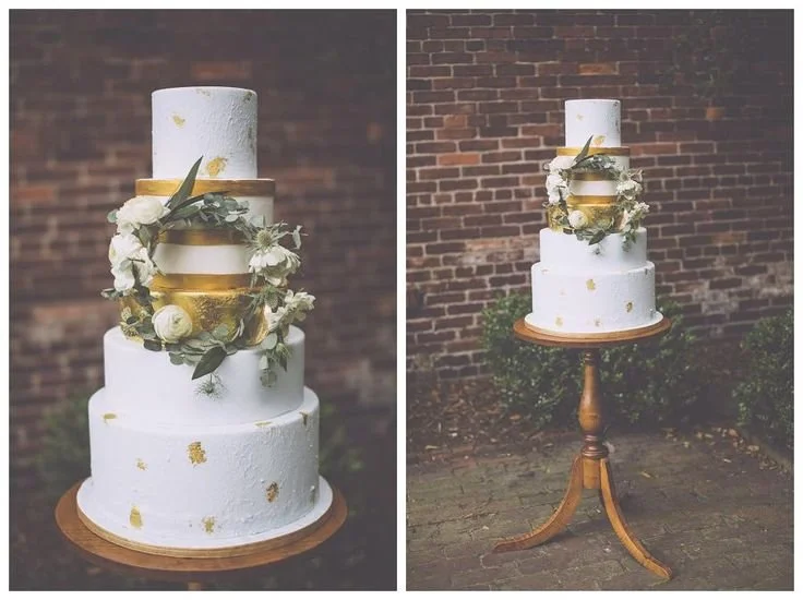 Three-tiered wedding cake with white frosting and gold accents, decorated with white flowers and greenery, displayed on a wooden cake stand outdoors against a brick wall.