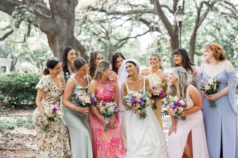 A group of women, including a bride in a white wedding dress and veil, and bridesmaids in pastel dresses, standing outdoors in a park with trees in the background, holding bouquets of flowers, smiling, and socializing.
