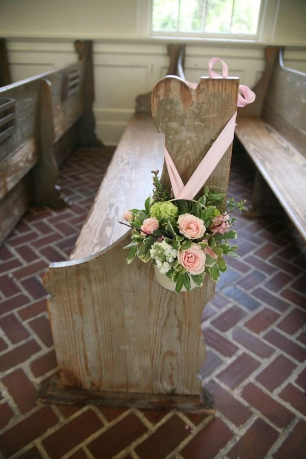 Wooden church pew with a heart-shaped piece of wood and a floral arrangement of pink roses and greenery attached to the front.