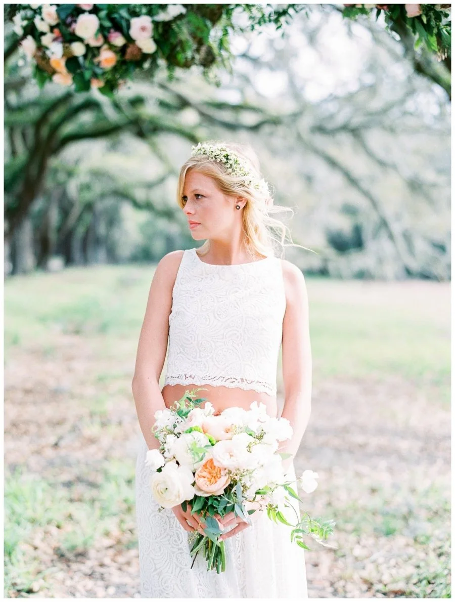 A young woman in a white lace dress holding a bouquet of flowers in an outdoor setting with trees and greenery.