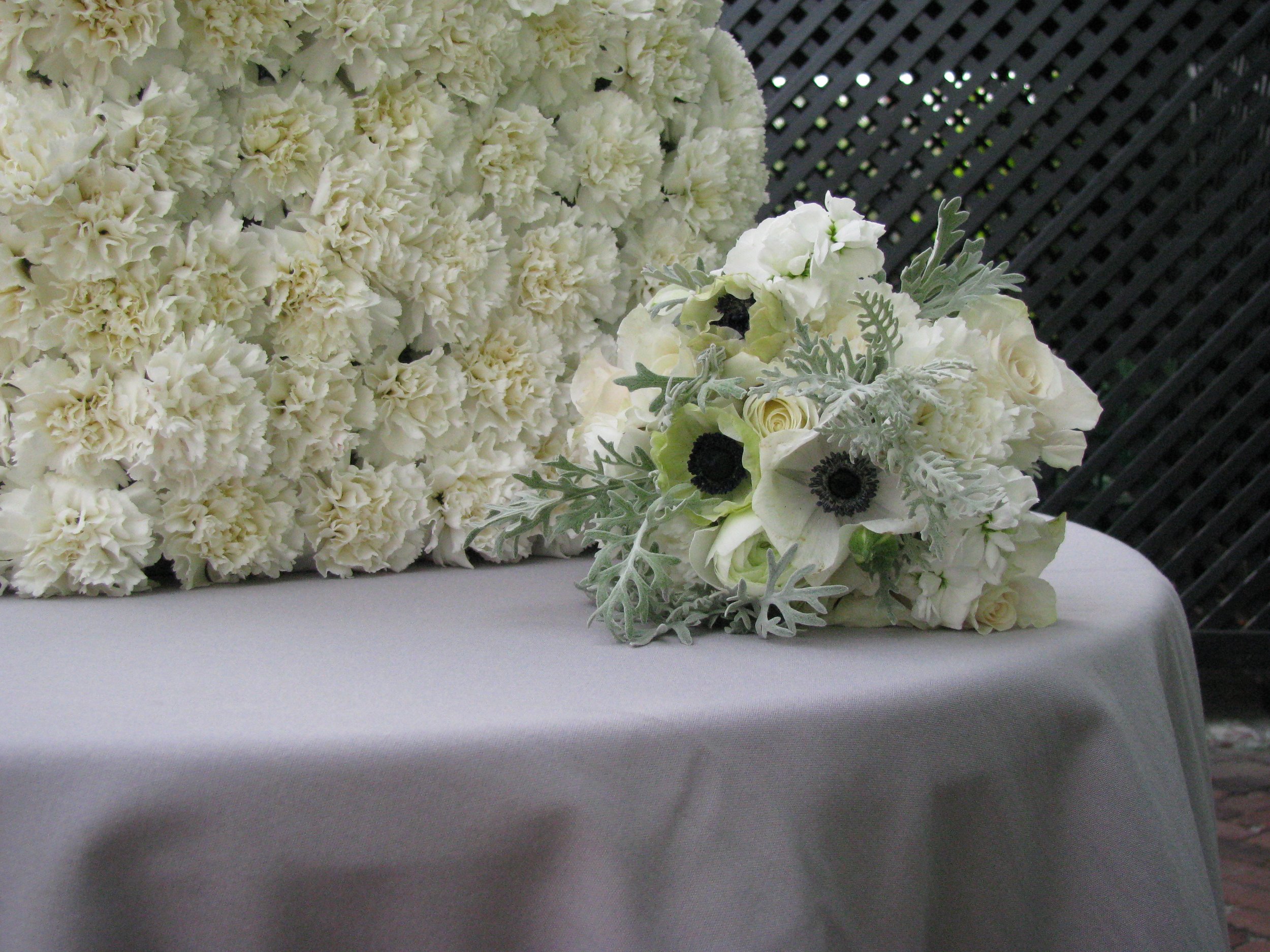 A large arrangement of white flowers, including carnations, roses, anemones, and dusty miller leaves, placed on a white tablecloth against a black lattice background.