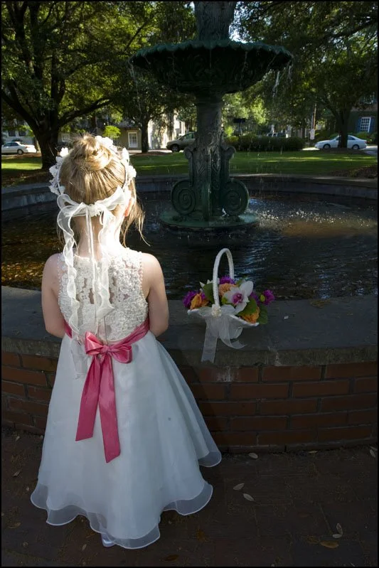 A young girl in a white dress with a pink ribbon standing in front of a decorative fountain with flowers in a park.