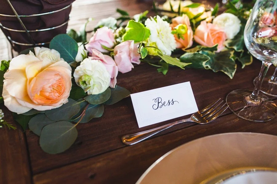 A wedding or event table setting decorated with a floral arrangement, a name card labeled 'Bess', a fork, and a wine glass.