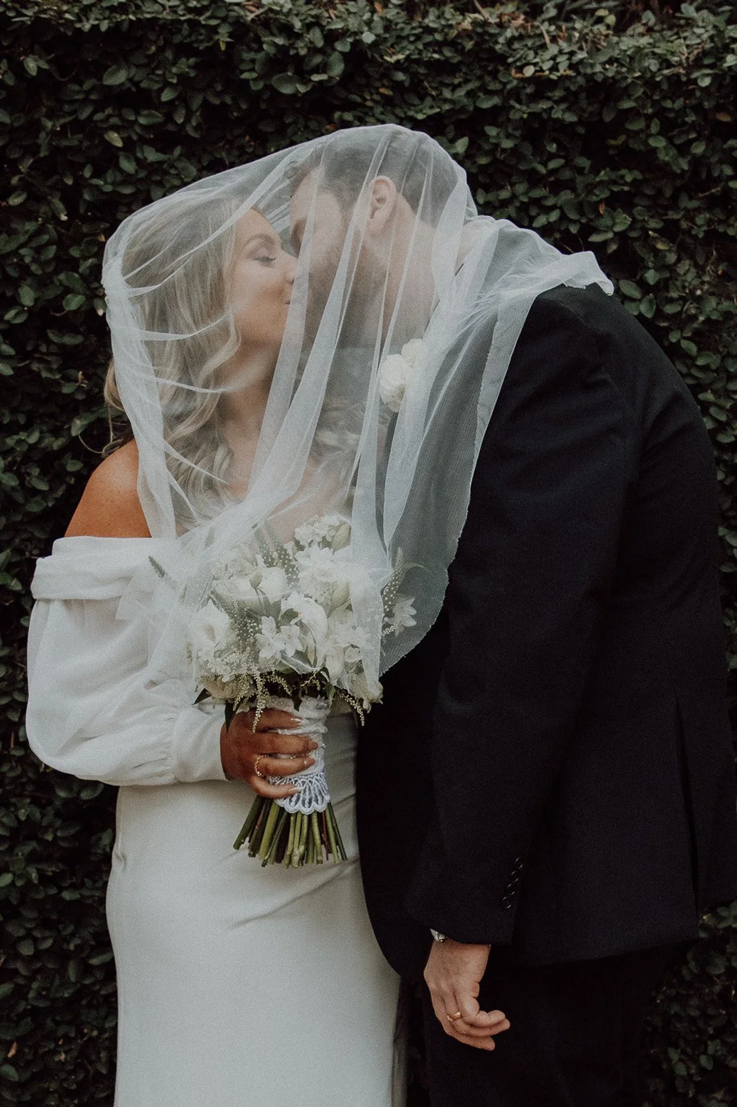 A bride and groom kiss under a sheer veil, with the bride holding a bouquet of white flowers, standing in front of a leafy green background.
