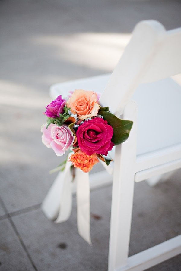 A white wooden chair decorated with a bouquet of colorful roses in pink, peach, orange, and purple, tied with a white ribbon.