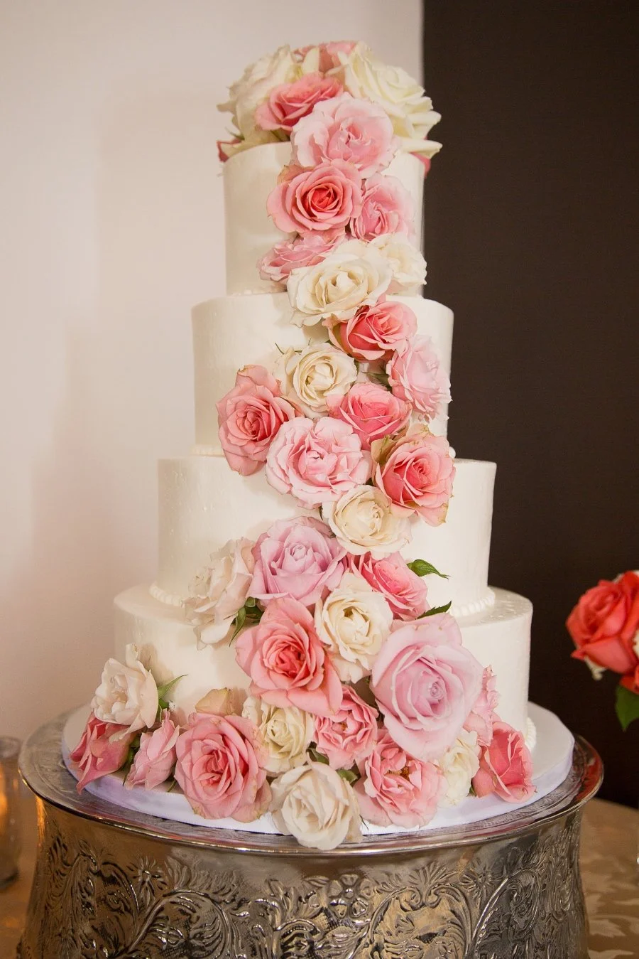 A four-tier white wedding cake decorated with cascading pink and white roses, placed on a silver ornate cake stand.