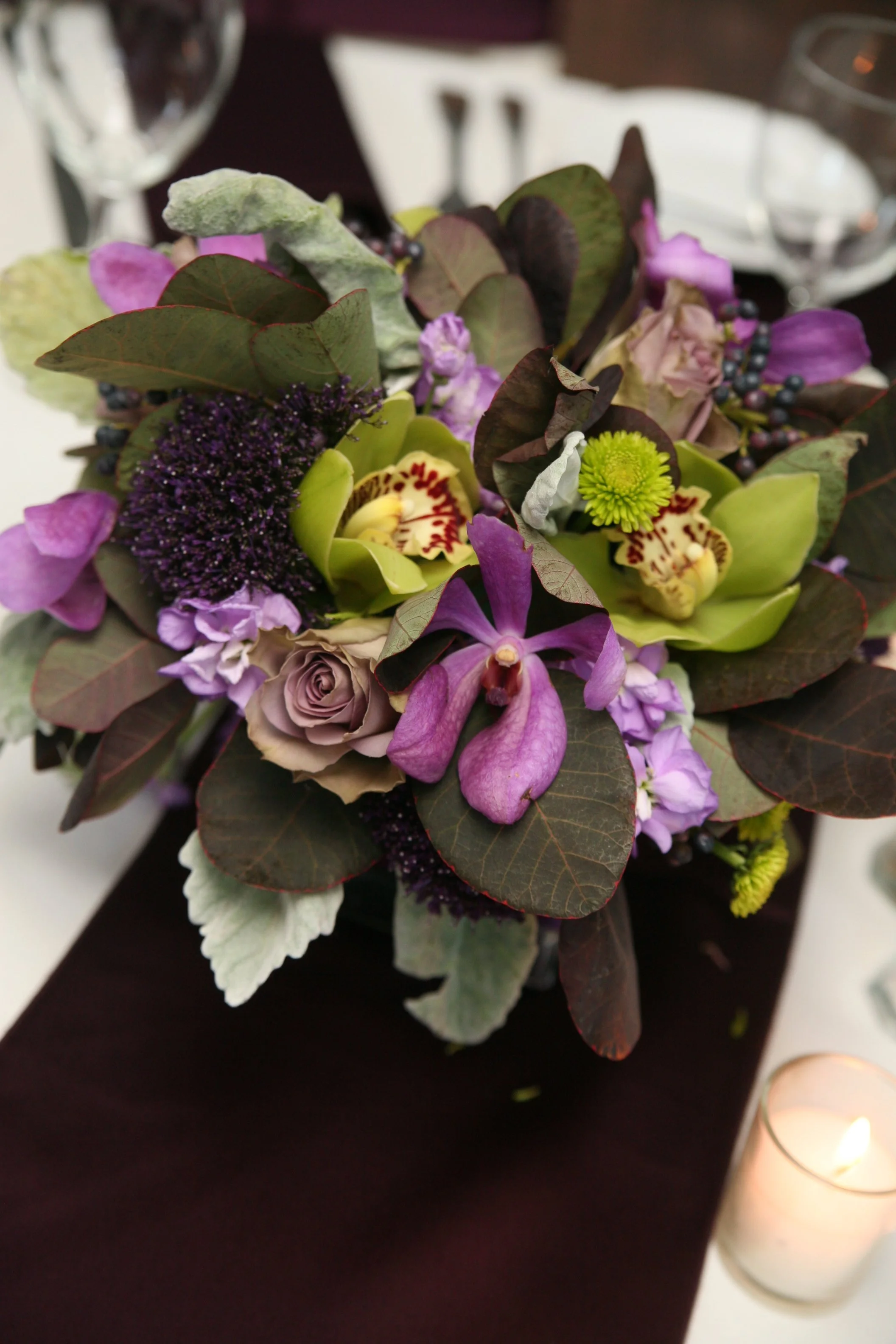A floral arrangement of purple and green flowers and leaves on a dark table with a candle and glassware in the background.