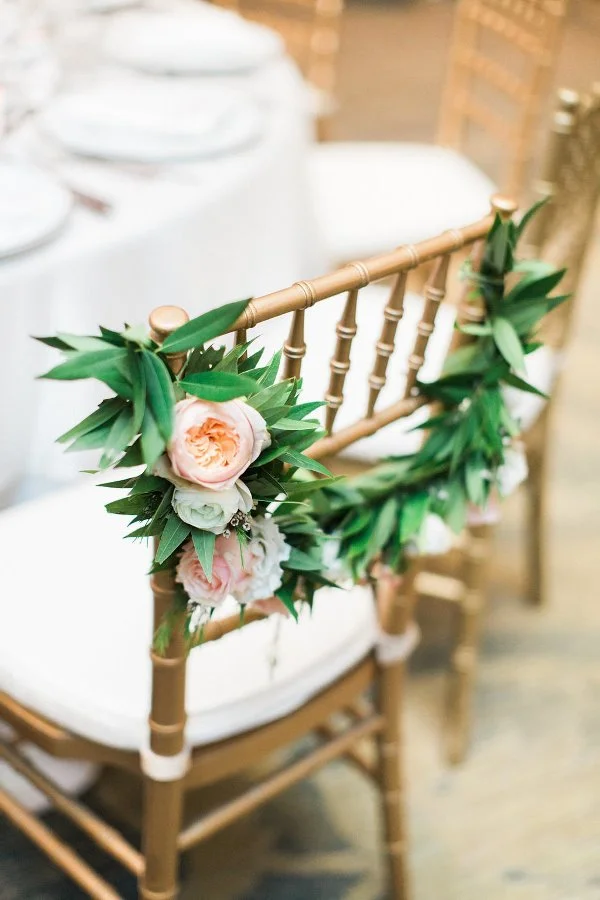 Gold Chiavari chair decorated with pink and white flowers and green leaves, set at a table with white tablecloths and place settings.