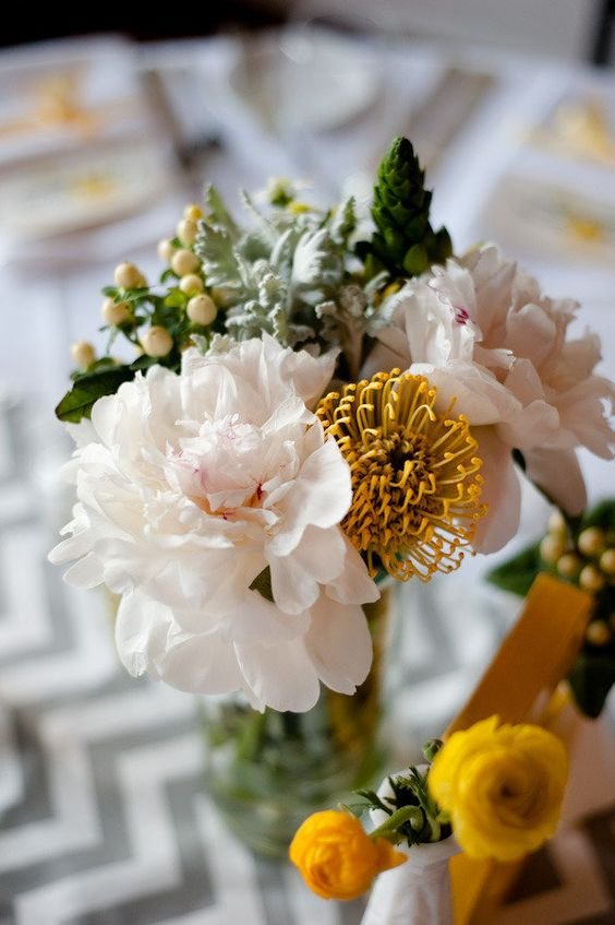 A bouquet of white peonies, yellow pincushion flowers, green buds, and foliage in a glass vase with a blurred patterned background.