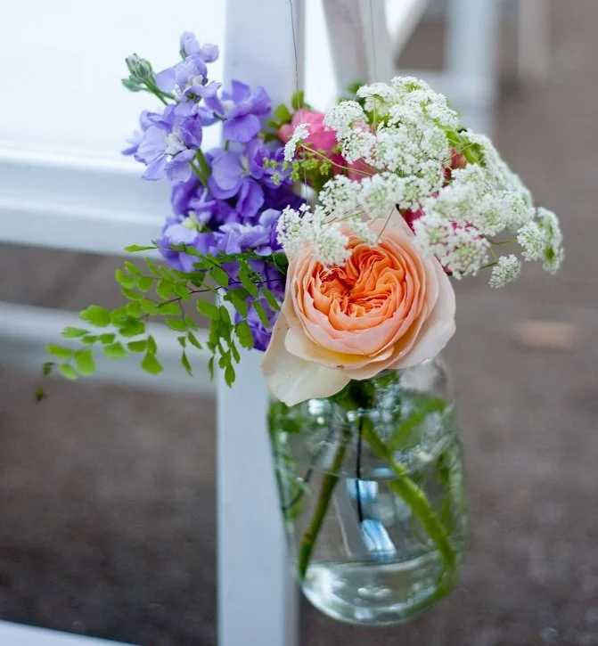 A glass jar with water holding a bouquet of pink and white roses, purple delphiniums, white baby's breath, and green foliage.