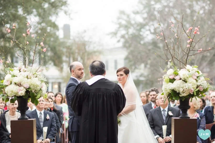 Wedding ceremony outdoors with bride and groom standing before officiant, surrounded by floral arrangements and guests seated in the background.
