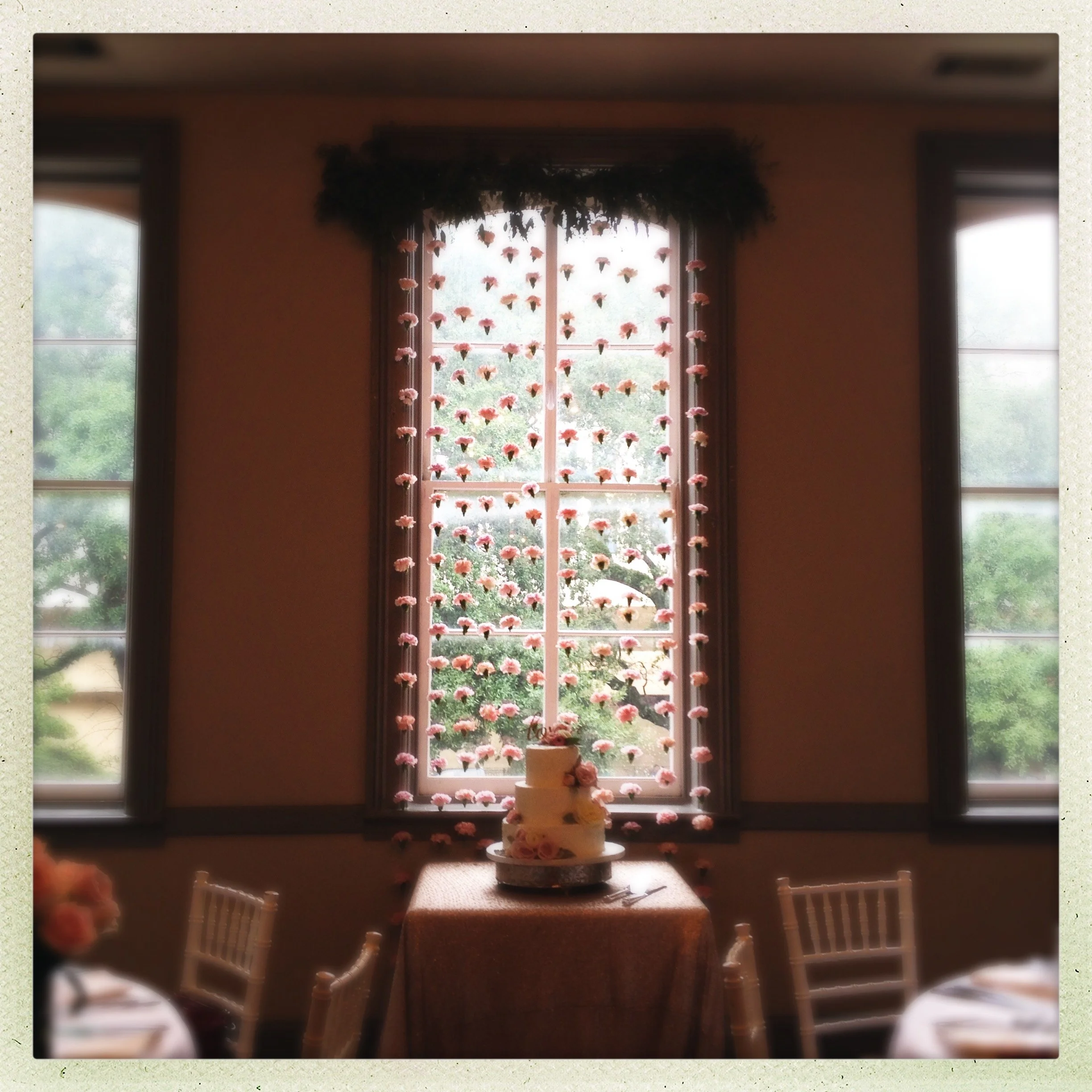 Decorated wedding cake on a table in front of a window with hanging pink flowers, next to chairs in a warmly lit room.