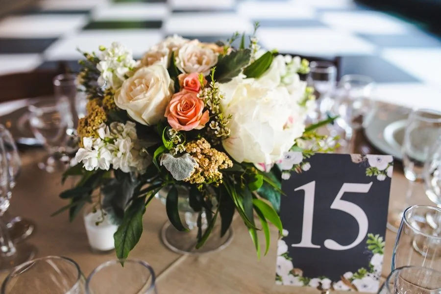 A floral centerpiece featuring white and blush roses, white hydrangeas, and greenery on a dinner table with clear glassware and a table number card reading 15.