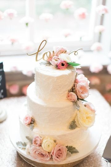 A three-tiered white wedding cake decorated with pink and white roses and a gold topper that says "love" on a table.