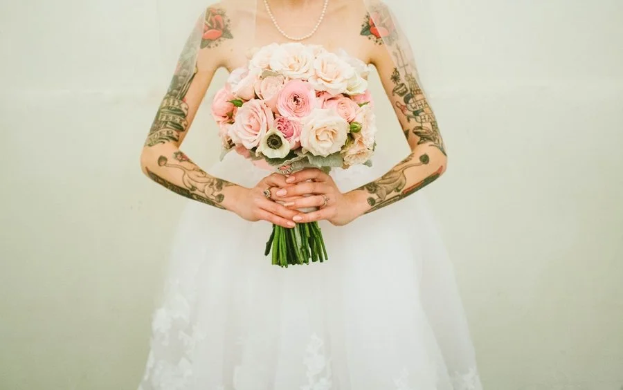 Bridal portrait showing a woman in a wedding dress holding a bouquet of pink and white roses, with tattoos on her arms visible.