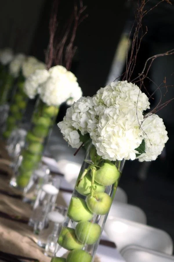 Vase with white hydrangea flowers and green apples inside, decorated with twigs, on a table at an event.