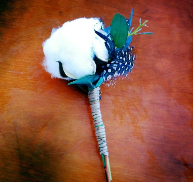 A cotton ball, feathers, leaves, and a stick wrapped in string arranged to look like a small decorative bouquet or flower on a wooden surface.