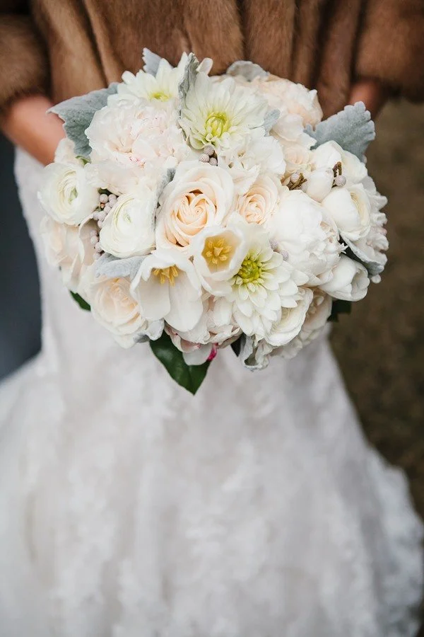 A bride holding a bouquet of white and pale pink flowers, including roses and dahlias, with green leaves against a blurred background.