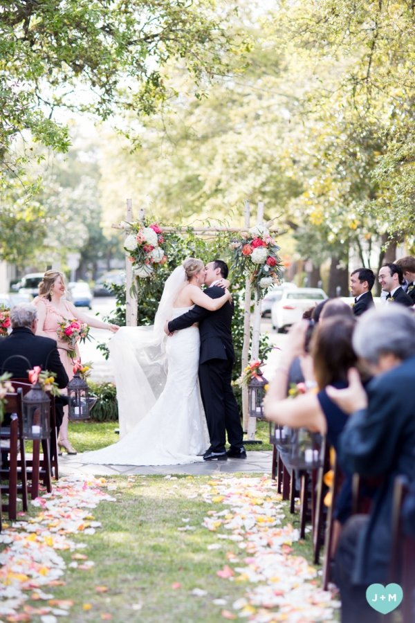 A bride and groom kiss during their outdoor wedding ceremony under a floral arch, with wedding guests seated on both sides, surrounded by trees and natural sunlight.