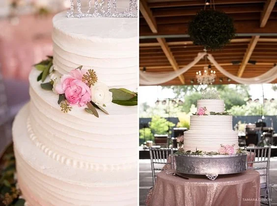 A three-tiered wedding cake decorated with pink and white flowers, topped with a silver 'Love' cake topper, set on a table at a reception hall with wooden beams, draped fabric, and a chandelier.