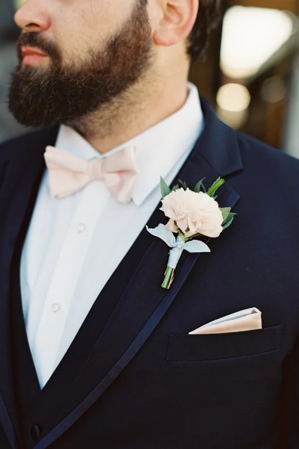 Close-up of a groom in a dark suit with a pink boutonniere, white shirt, and pink bow tie.