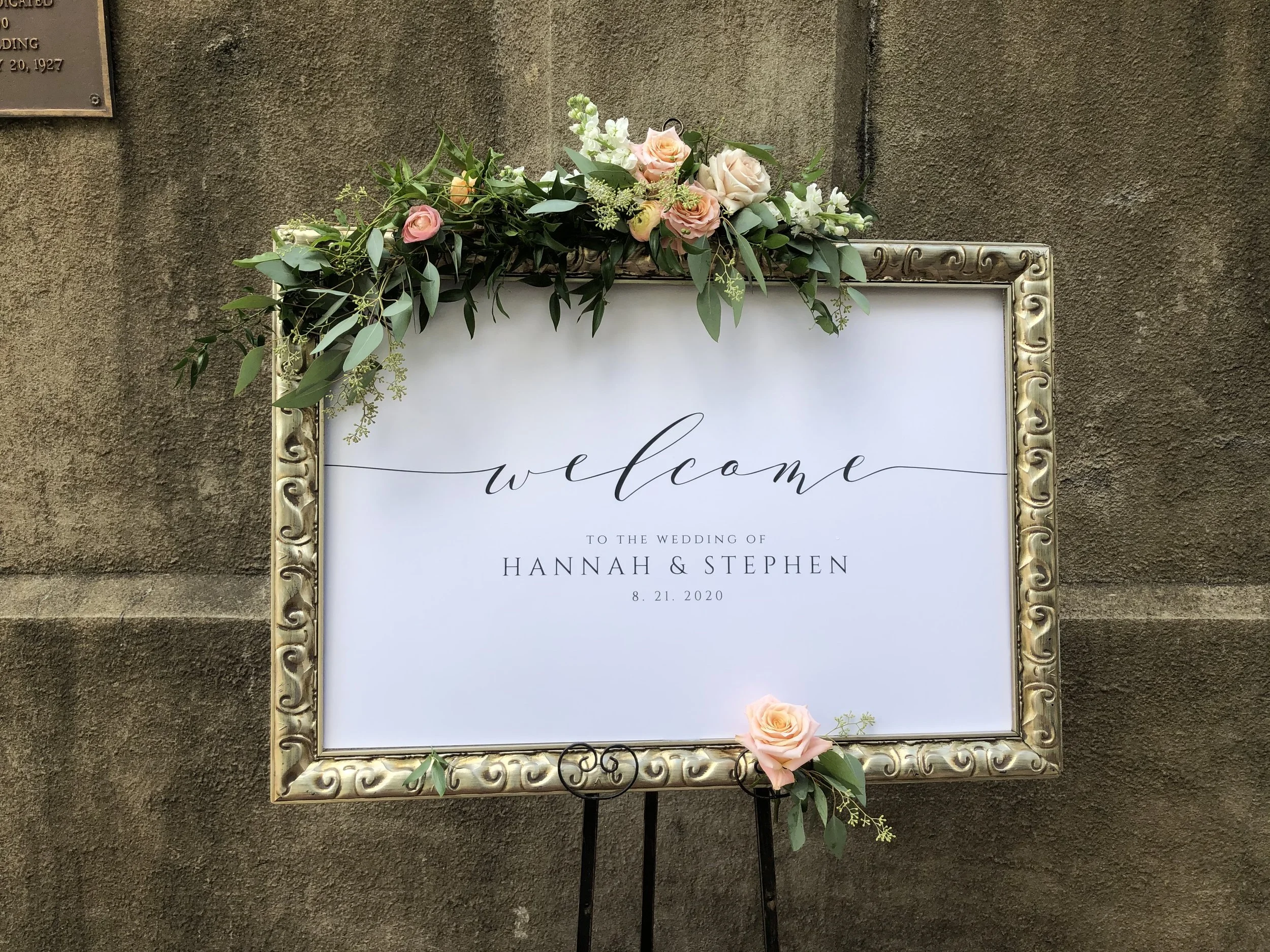 Wedding welcome sign decorated with pink and white roses, and green foliage, placed on a stand against a textured wall, for the wedding of Hannah and Stephen on August 21, 2020.