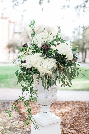 Elegant floral arrangement with white hydrangeas and green foliage in a white vase outside on a pedestal.