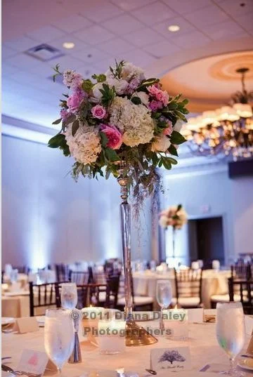 Elegant table setting with a tall floral centerpiece in a banquet hall, featuring pink and white flowers, wine glasses, and place cards.