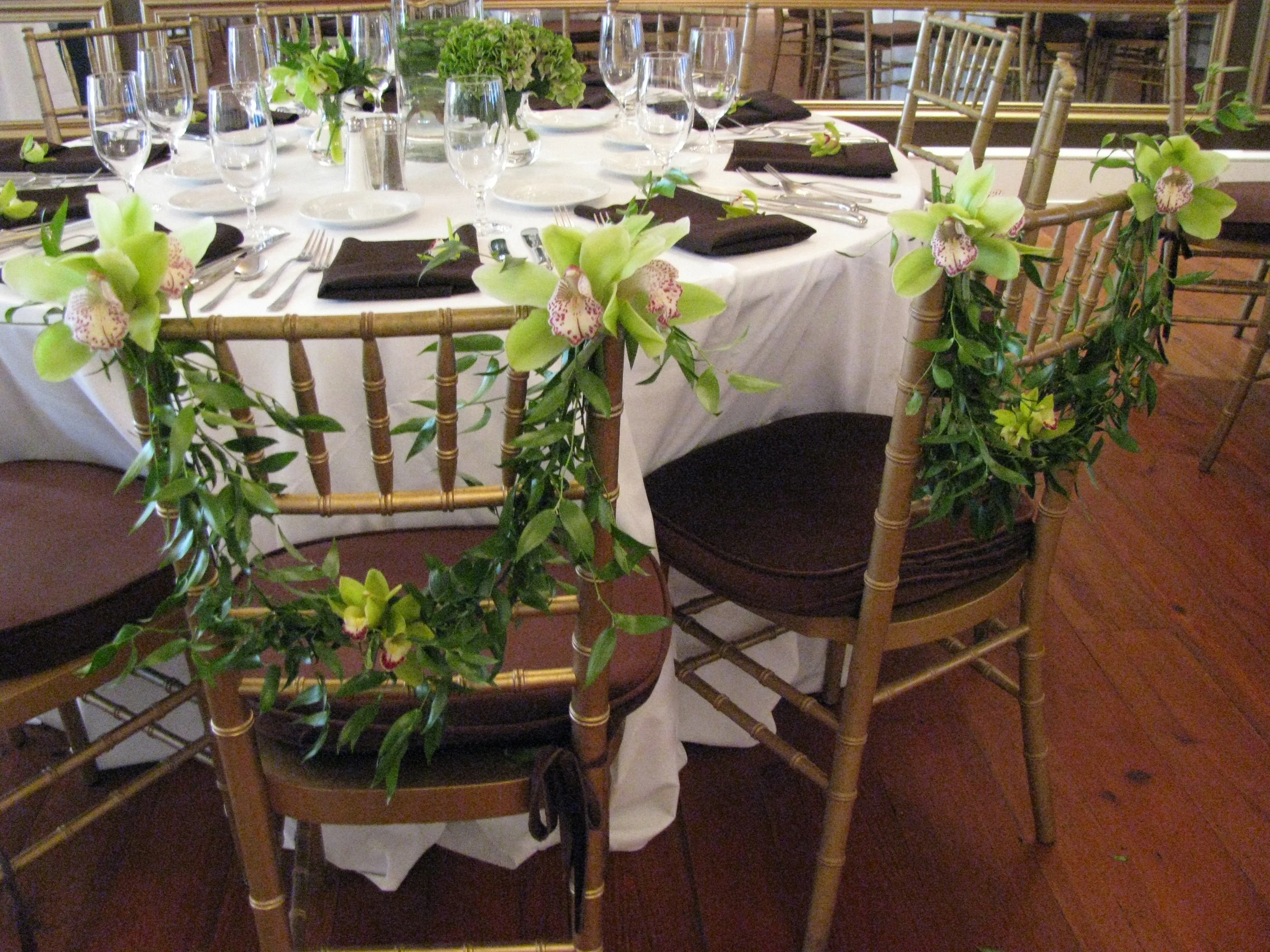A round banquet table with a white tablecloth, set with glassware, black napkins, and silverware, decorated with pink and green orchids and greenery, surrounded by gold-colored chairs with brown cushions.