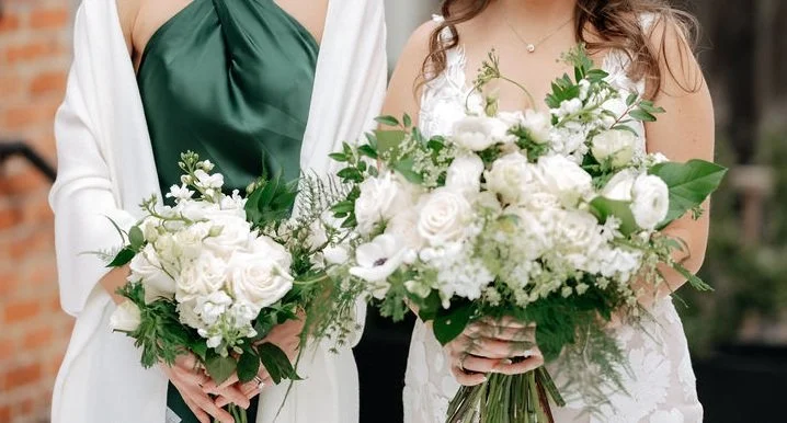 Two women in wedding dresses holding large bouquets of white flowers and greenery.