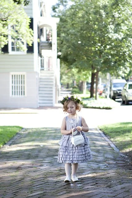 A young girl wearing a gray frilly dress, white shoes, and a floral headband walking along a brick sidewalk while holding a small white purse.