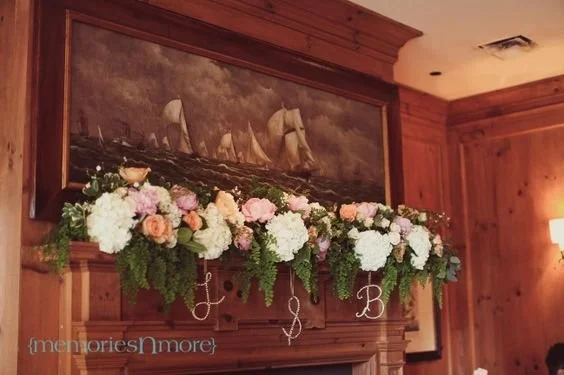 A wooden fireplace mantel decorated with a floral arrangement of white and pink flowers and greenery, with a framed painting of sailing ships above it.