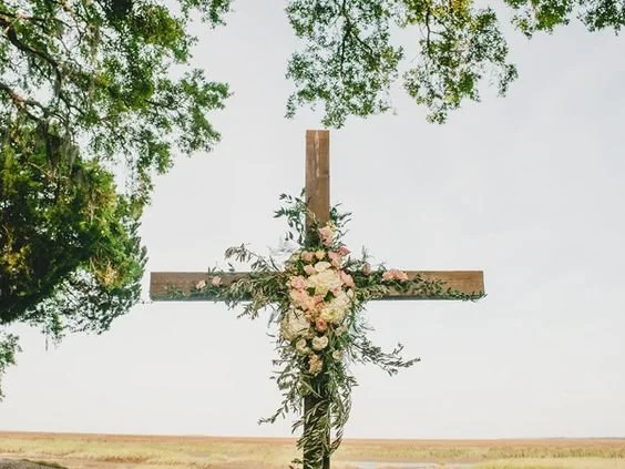 A wooden cross decorated with pink and white flowers standing outdoors in a field with trees around.
