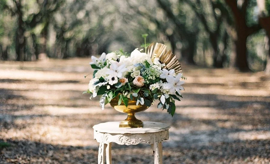 A floral arrangement in a gold vase on an ornate white pedestal table, set outdoors on a dirt path with trees in the background.