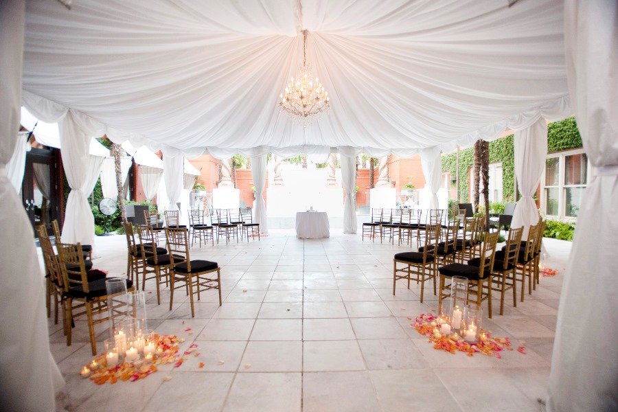 Empty wedding ceremony setup inside a white tent with elegant draped fabric, a chandelier, gold chairs with black cushions arranged in two rows, and candle-lit flower petal decor on the floor.