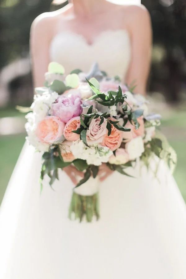 Bride holding a bouquet of pink and white flowers in a white wedding dress outdoor setting.