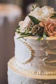 A wedding cake decorated with white and blush roses, greenery, and a pearl necklace.