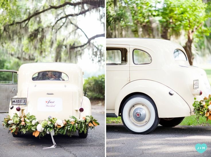A vintage cream-colored car decorated with flowers for a wedding, with a 'Just Married' sign, parked outdoors surrounded by green trees.