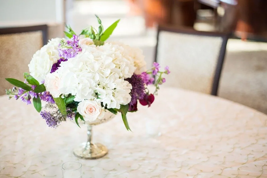 Elegant centerpiece with white hydrangeas, roses, and purple flowers on a table with a cream-colored tablecloth and chairs in the background.