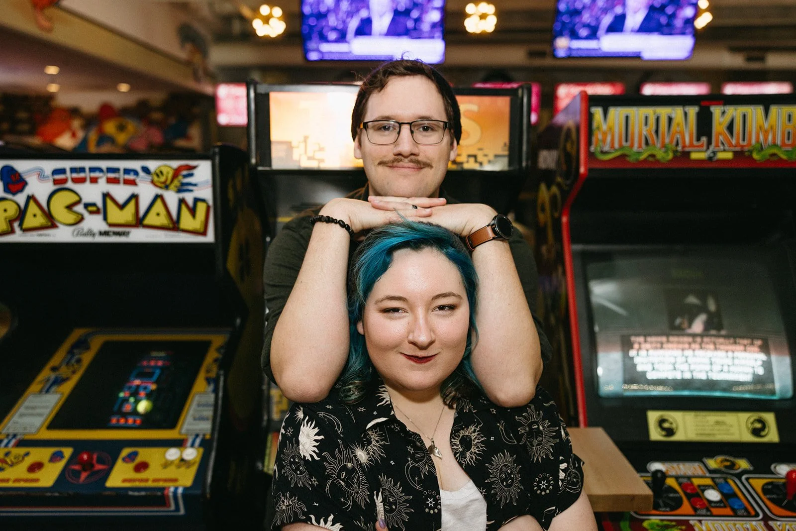 Two people, one with blue hair and one with glasses and dark hair, are posing in front of arcade machines, including Pac-Man and Mortal Kombat, in an arcade. Mackie and Connor Nagelkirk