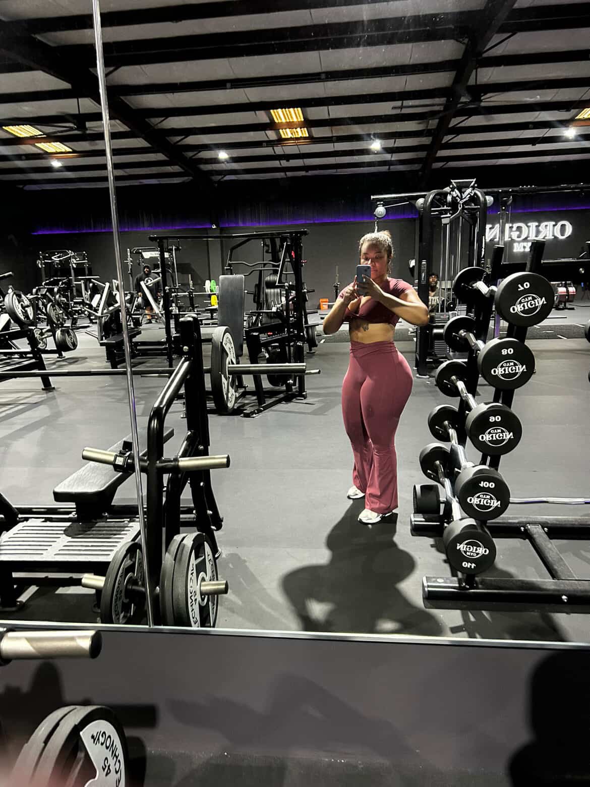 Woman taking a mirror selfie at a gym, wearing pink workout clothes, surrounded by weightlifting equipment and mirrors.