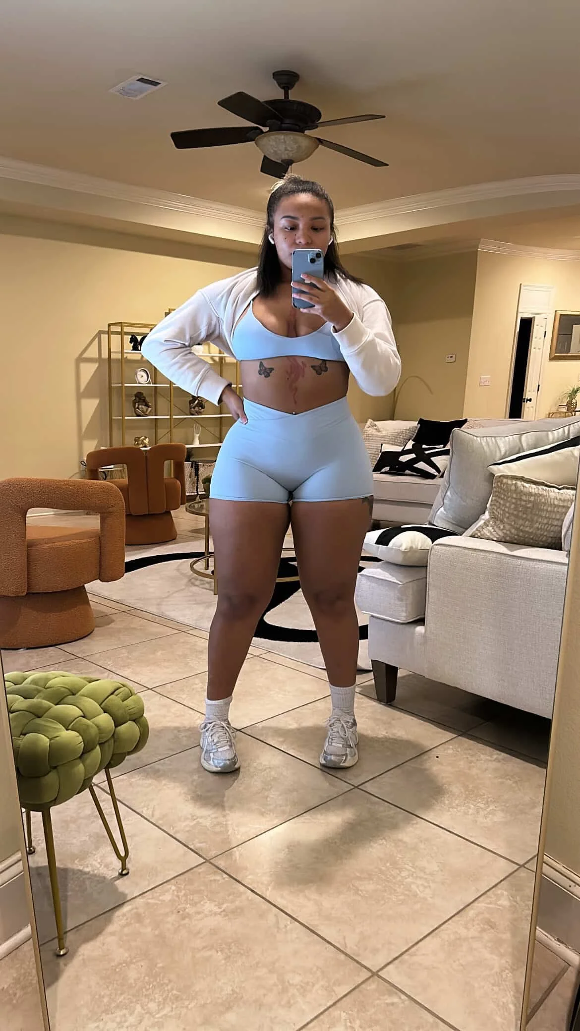 A woman taking a mirror selfie in a living room, wearing workout clothes including a sports bra, shorts, and sneakers.