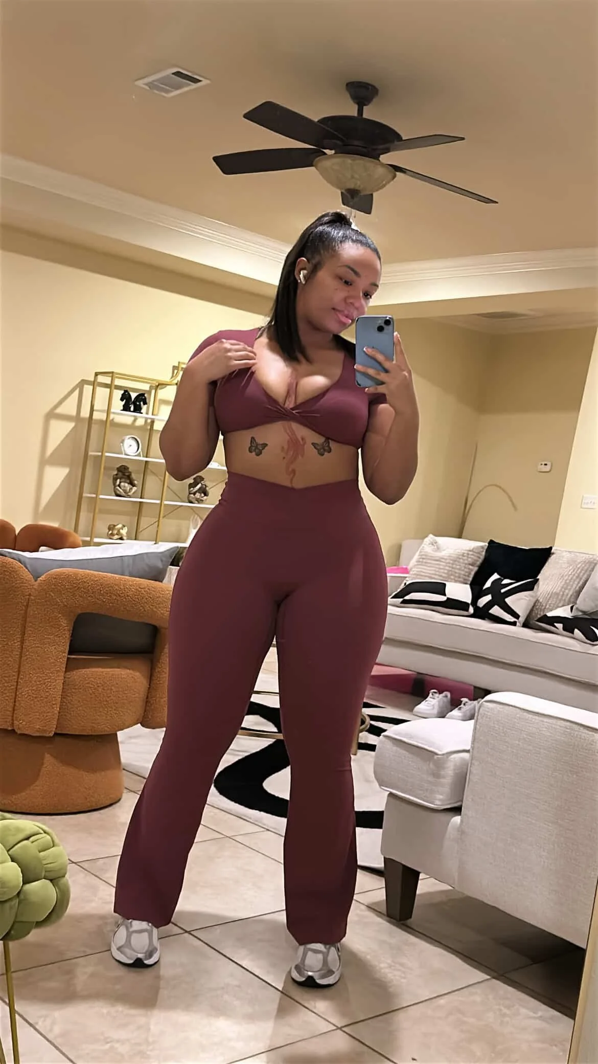 A woman taking a mirror selfie in a living room, dressed in a matching maroon workout set with butterflies and flame tattoos visible on her torso, wearing sneakers, with a ceiling fan and modern decor in the background.