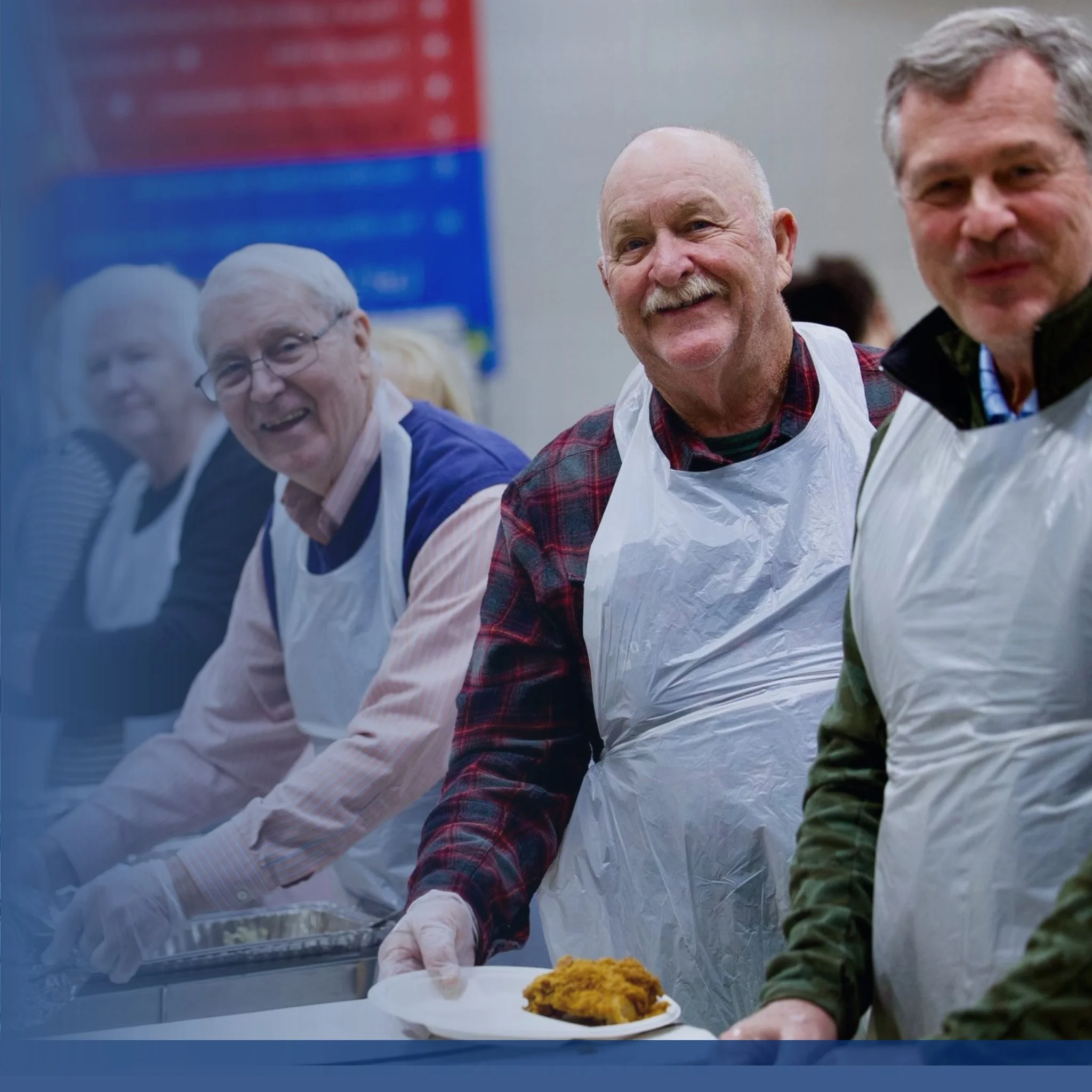 A group of people, likely volunteers, are serving food at a community event. One person is placing a piece of cornbread on a white plate, and they are wearing gloves. The background includes colorful banners or posters.