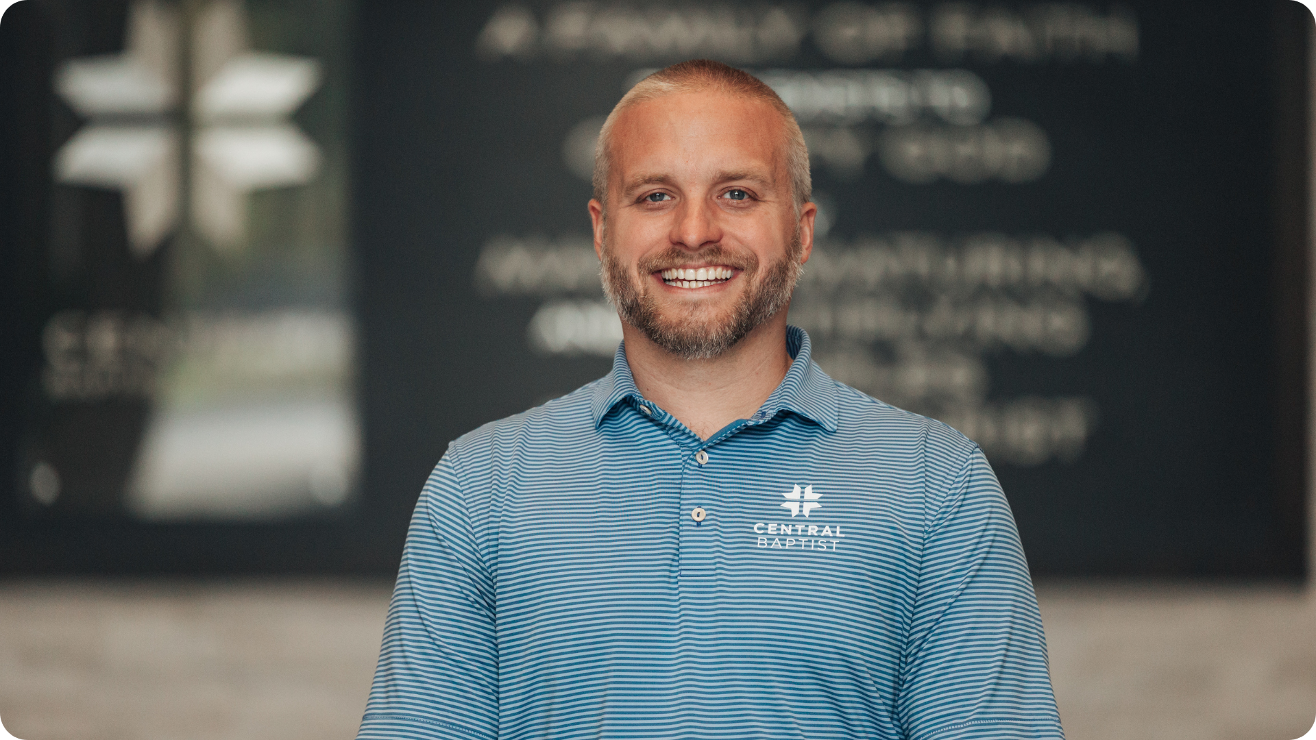 A smiling man with a beard and short blonde hair wearing a blue striped polo shirt with the logo of Central Baptist church, standing indoors with a blurred background.