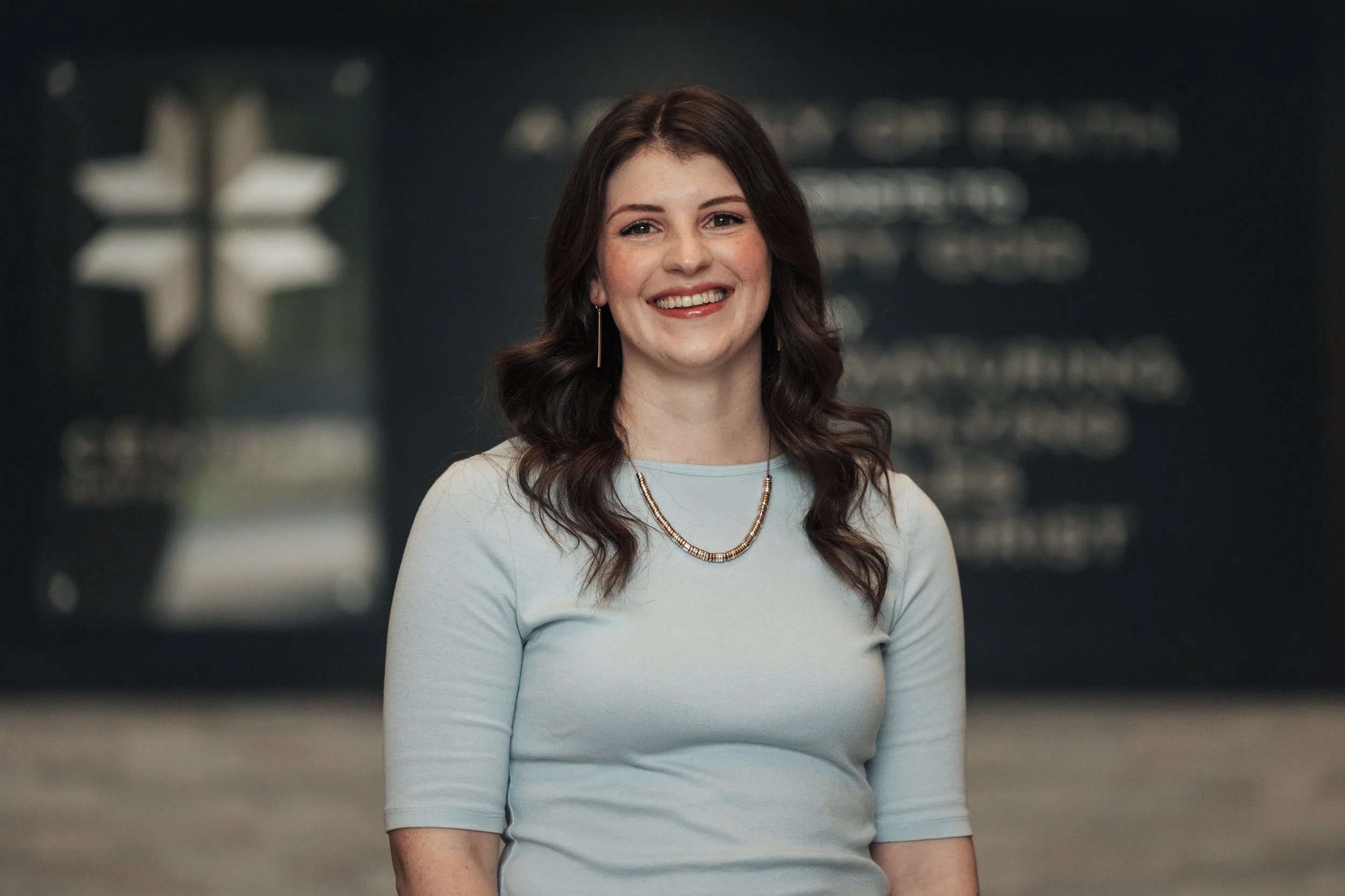 A woman with shoulder-length dark brown hair, smiling, wearing a light blue top, a beaded necklace, and earrings, standing in front of a blurred dark background with some white lights and text.