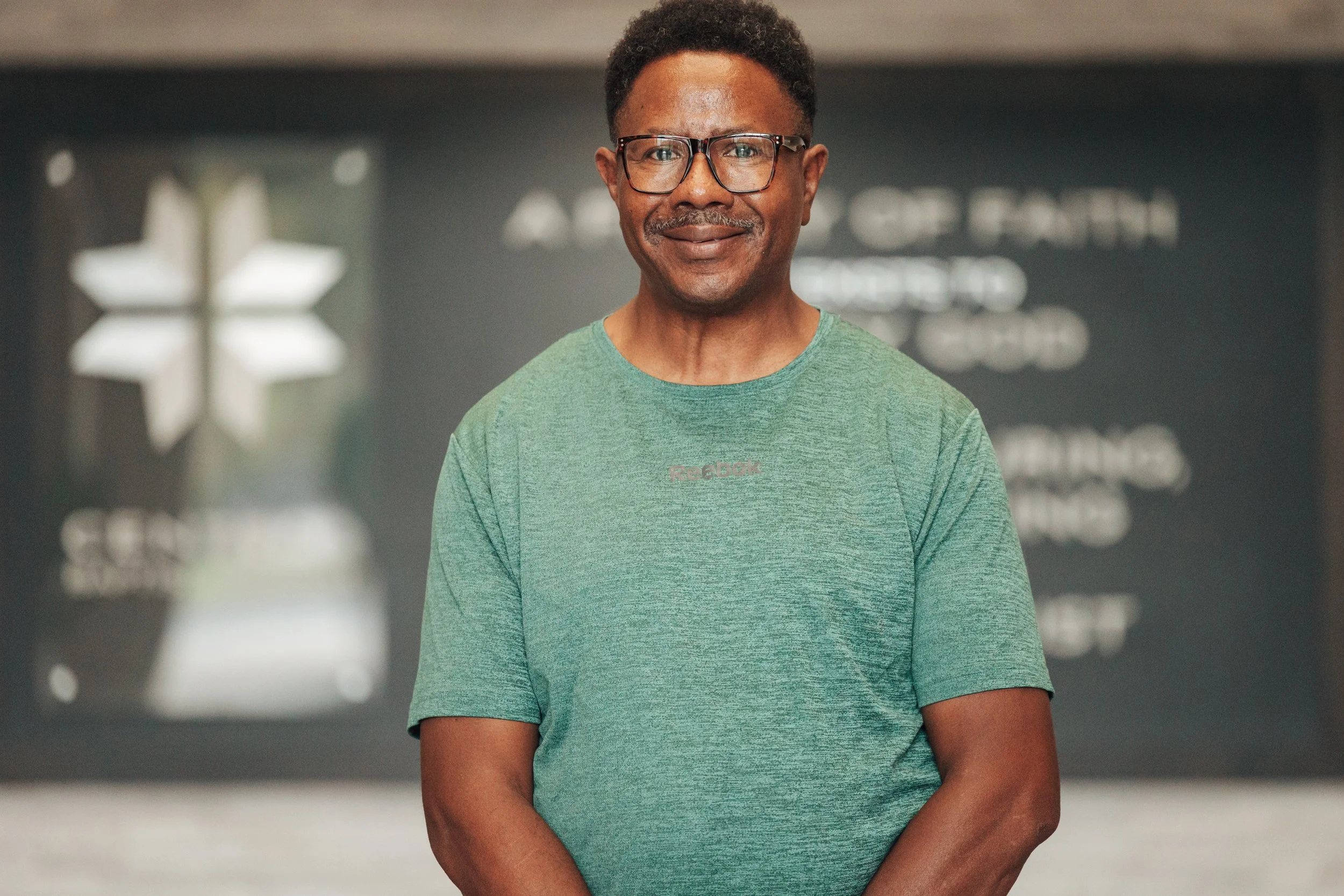 A middle-aged man with short curly hair, glasses, and a light green Reebok T-shirt standing indoors with a slight smile.