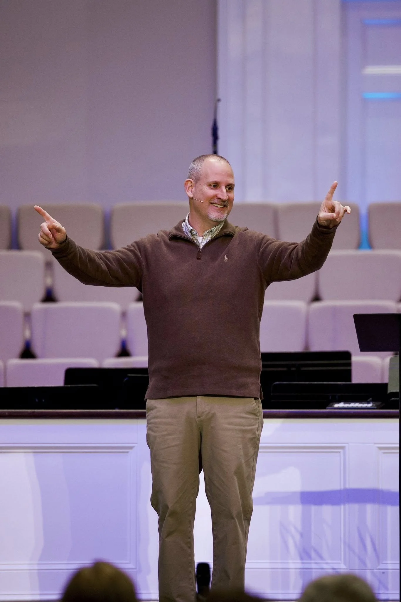 A middle-aged man with a beard, wearing a brown pullover and khaki pants, standing in front of a stage, smiling, and gesturing with both hands pointing outward.